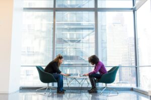 Two women engage in remote work on laptops in a bright, modern office lobby, reflecting a professional yet relaxed atmosphere.